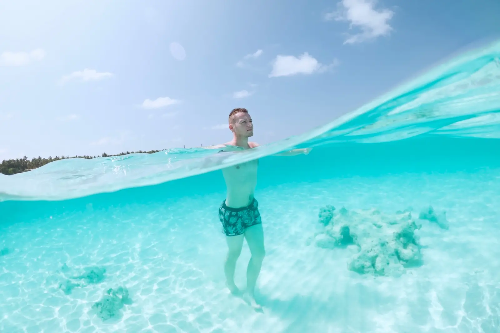 Split view of a man standing in clear turquoise shallows above sand and coral patches