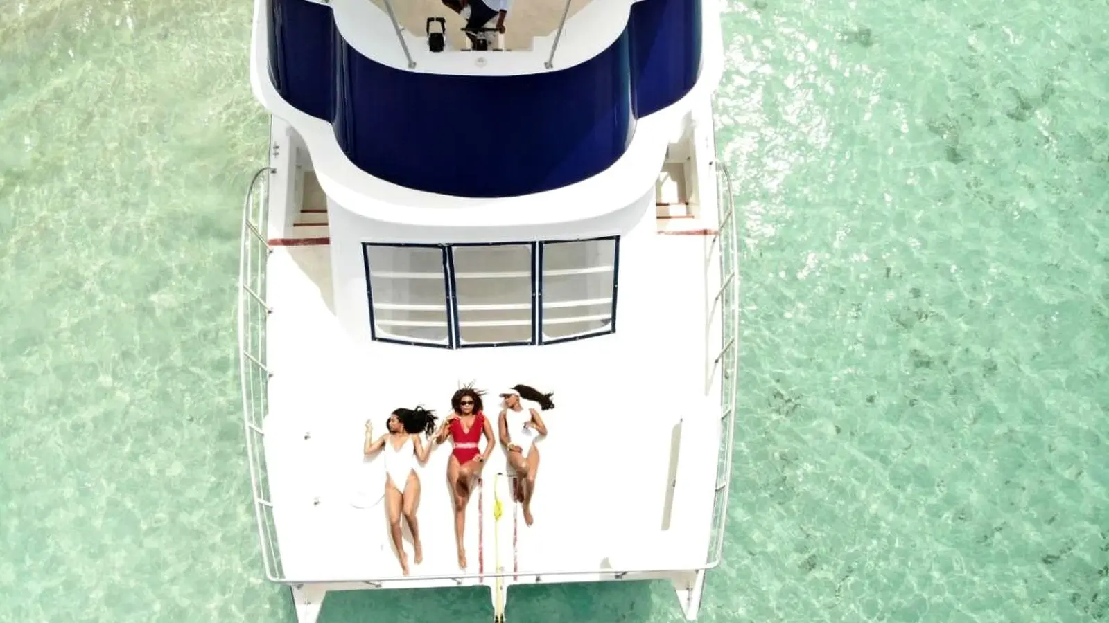 Overhead view of three women sunbathing on the bow of a power catamaran over pale green shallows