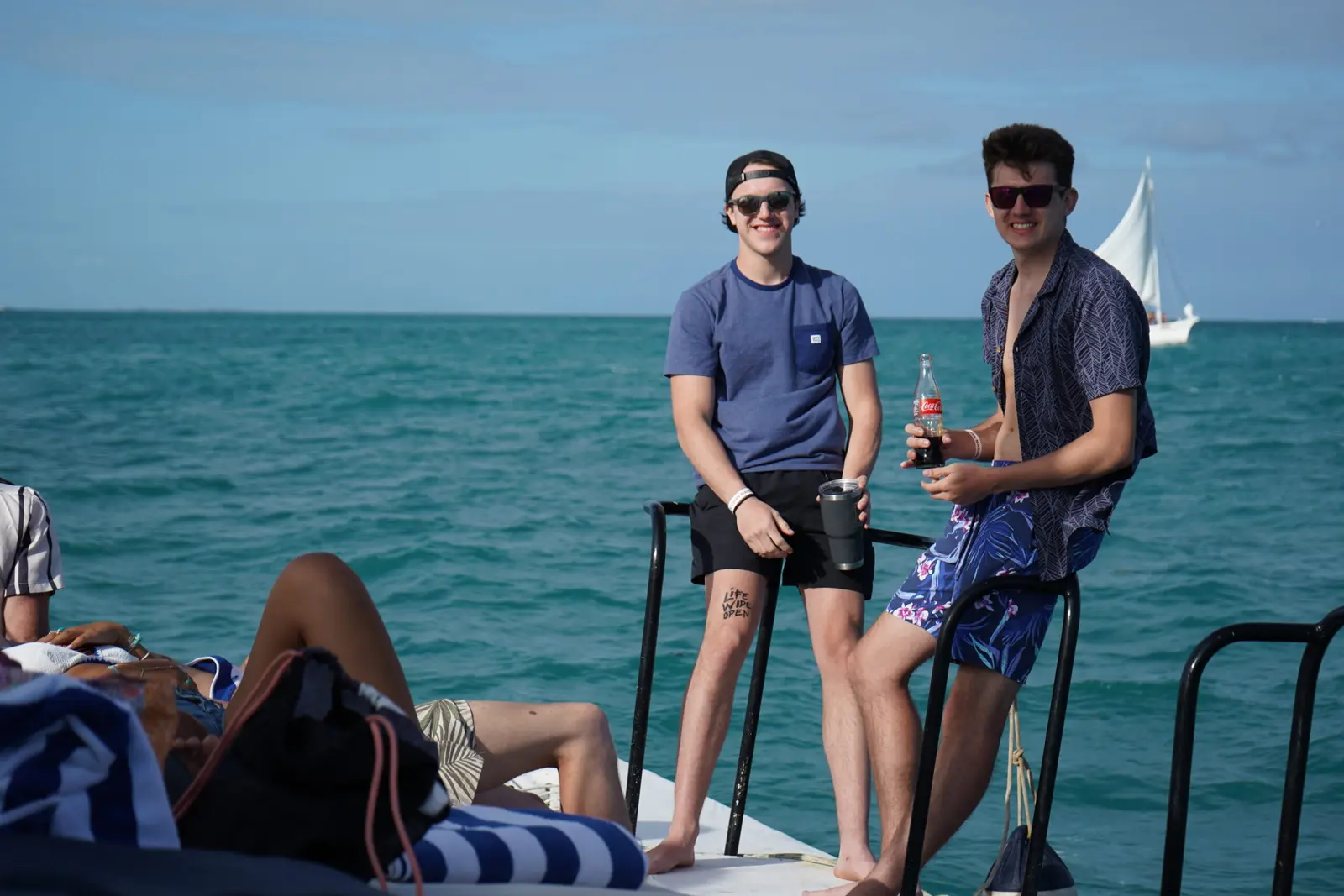 Two men with drinks leaning on the catamaran rail off Belize, a sailboat on the horizon behind
