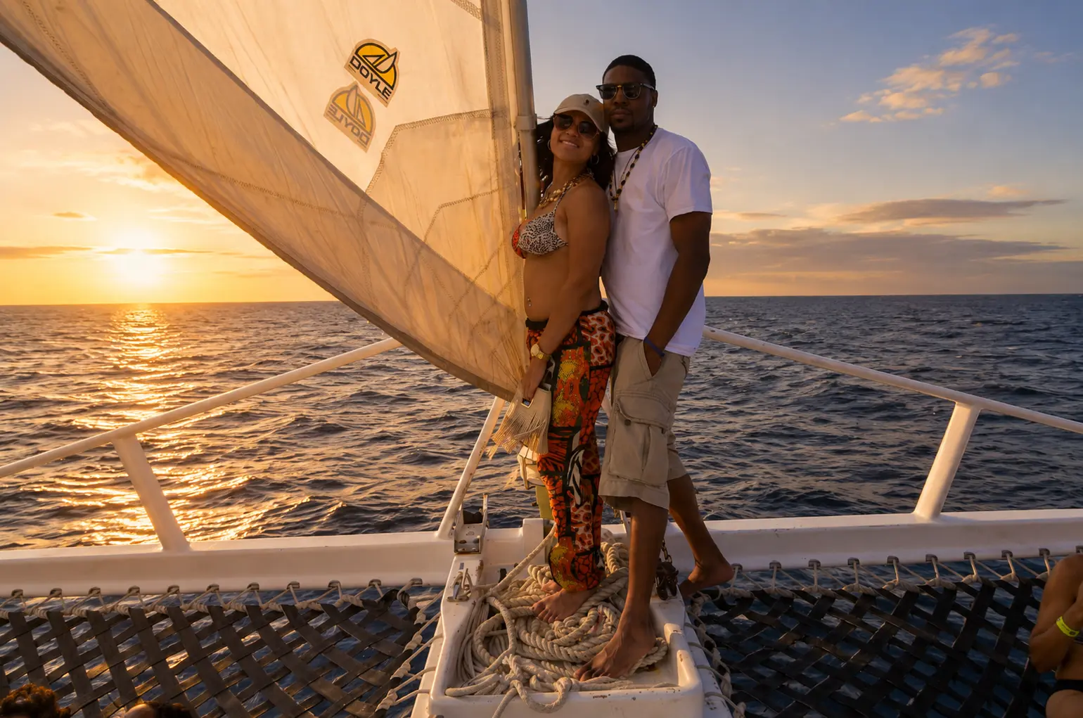 Couple posing beside the sail on the bow net of a sailing catamaran at sunset off Montego Bay