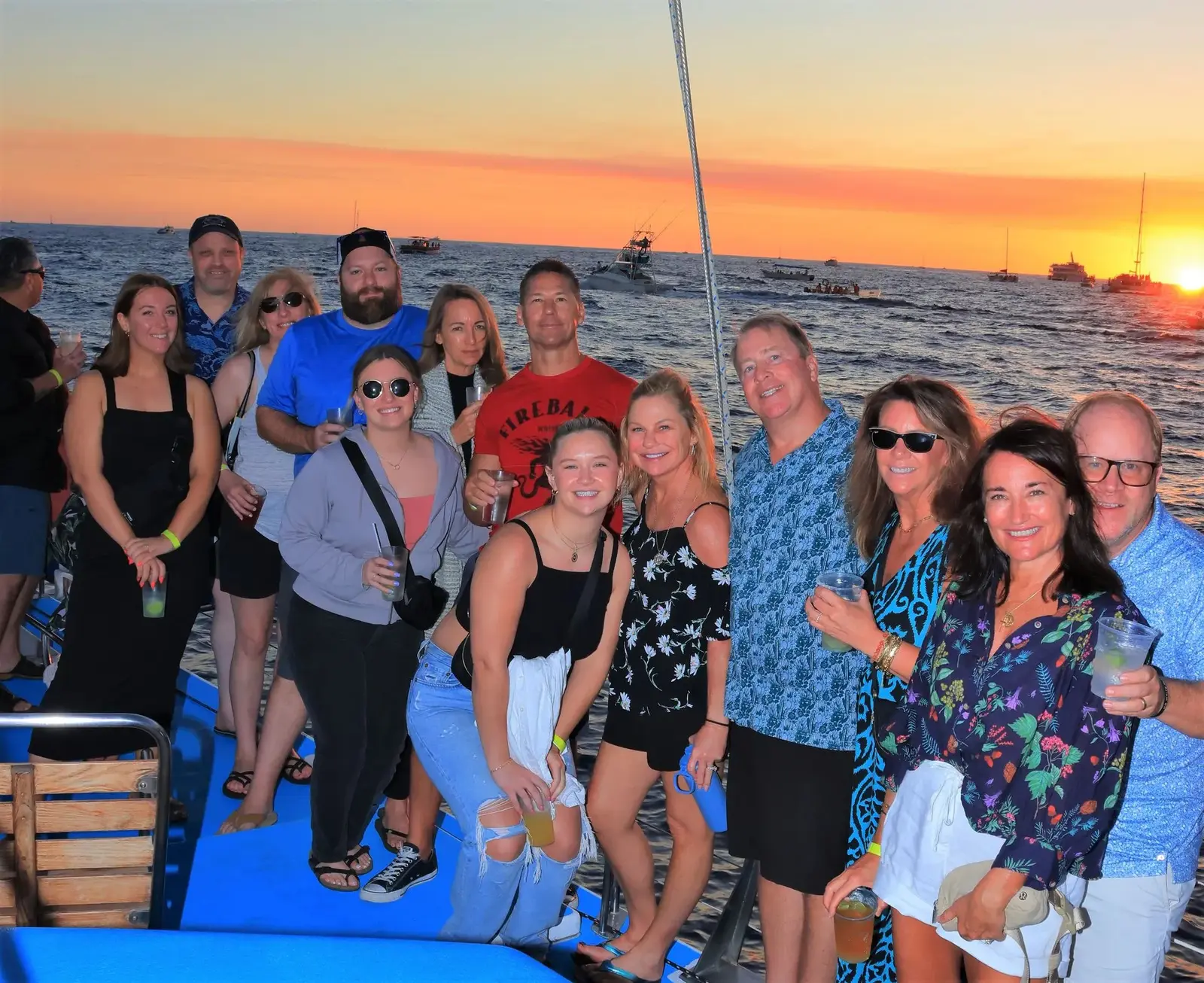 Large group of guests holding drinks on a catamaran deck at sunset off Cabo San Lucas