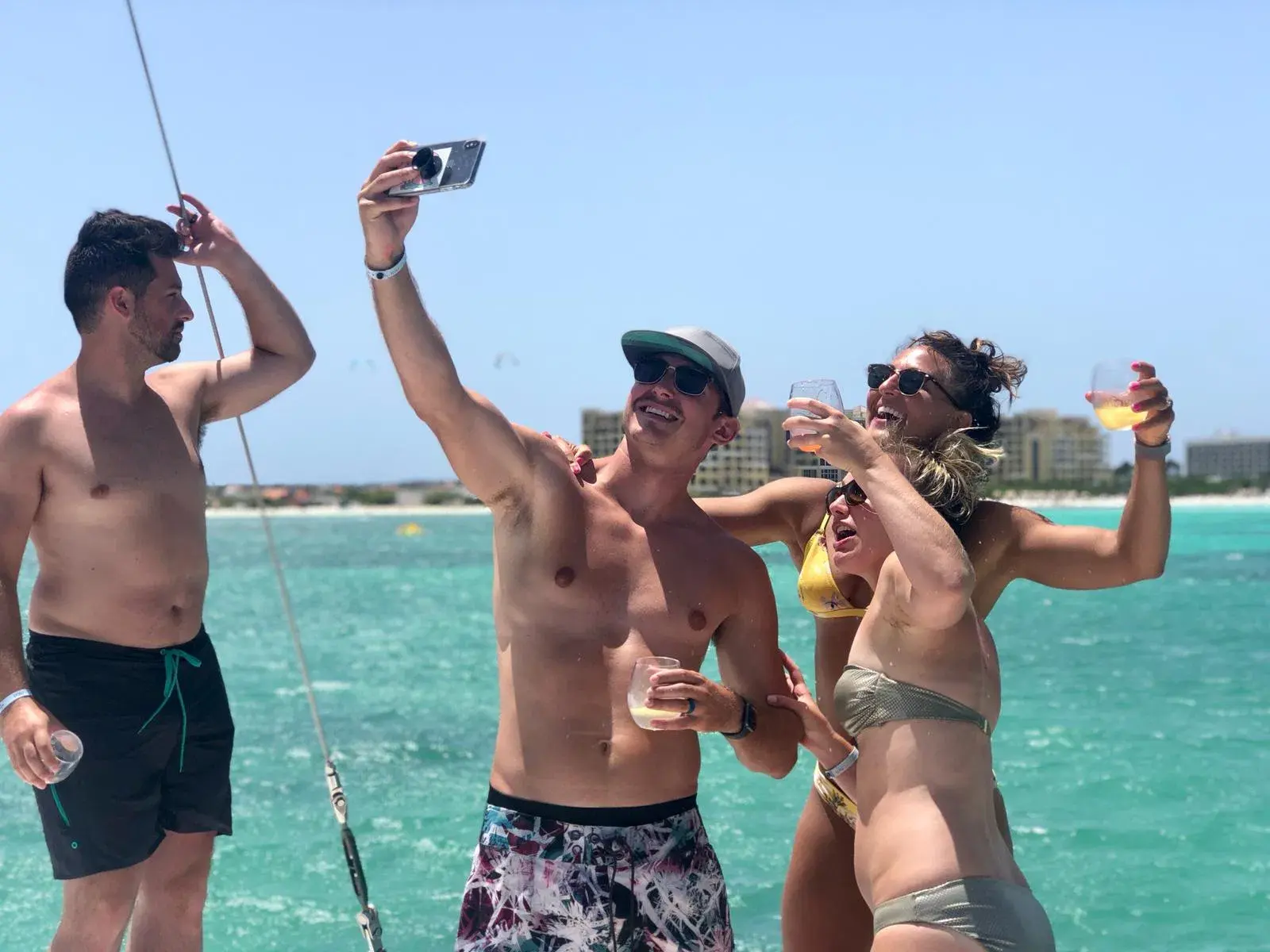 Four guests taking a selfie with drinks on deck, Aruba's turquoise water and hotels behind