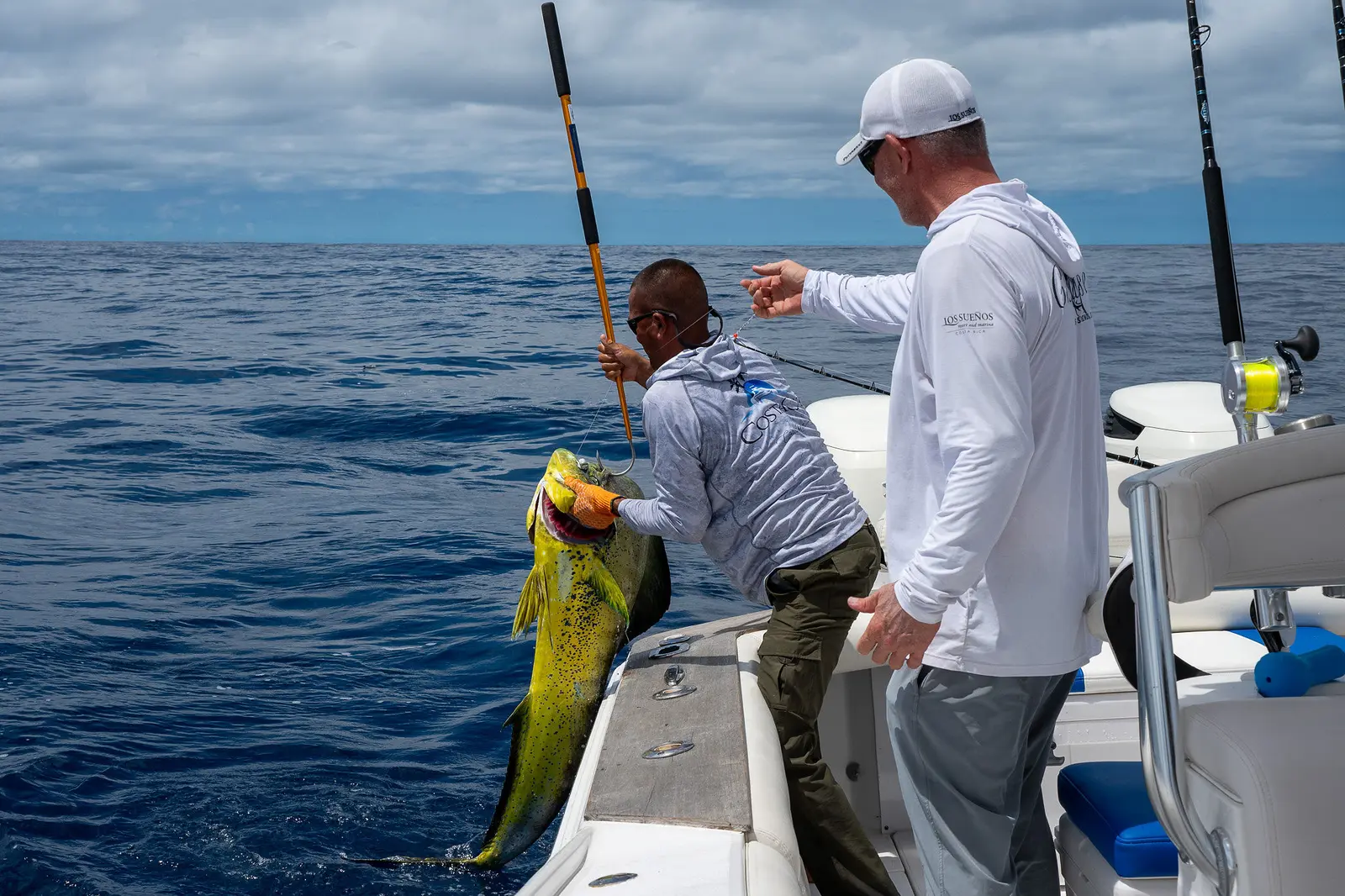 Crew member gaffing a green and gold mahi-mahi over the side of a center-console boat as an angler watches