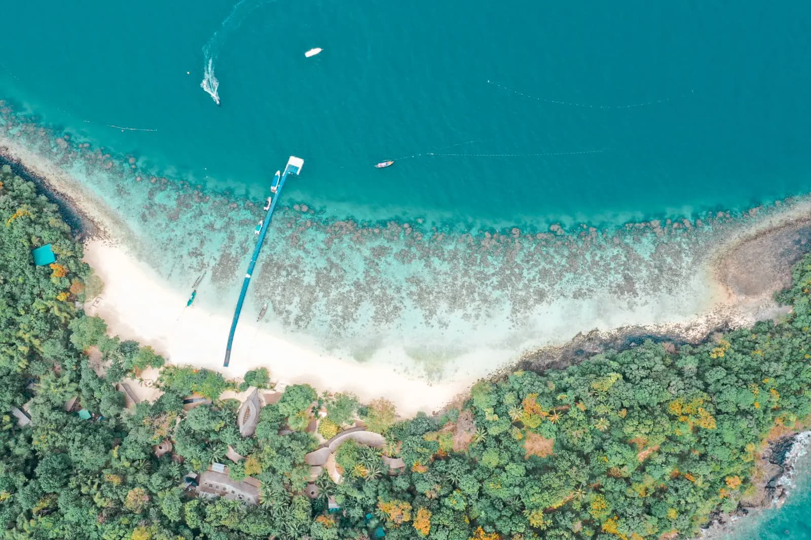 Aerial view of a long jetty and white-sand bay curving around a forested island in Thailand