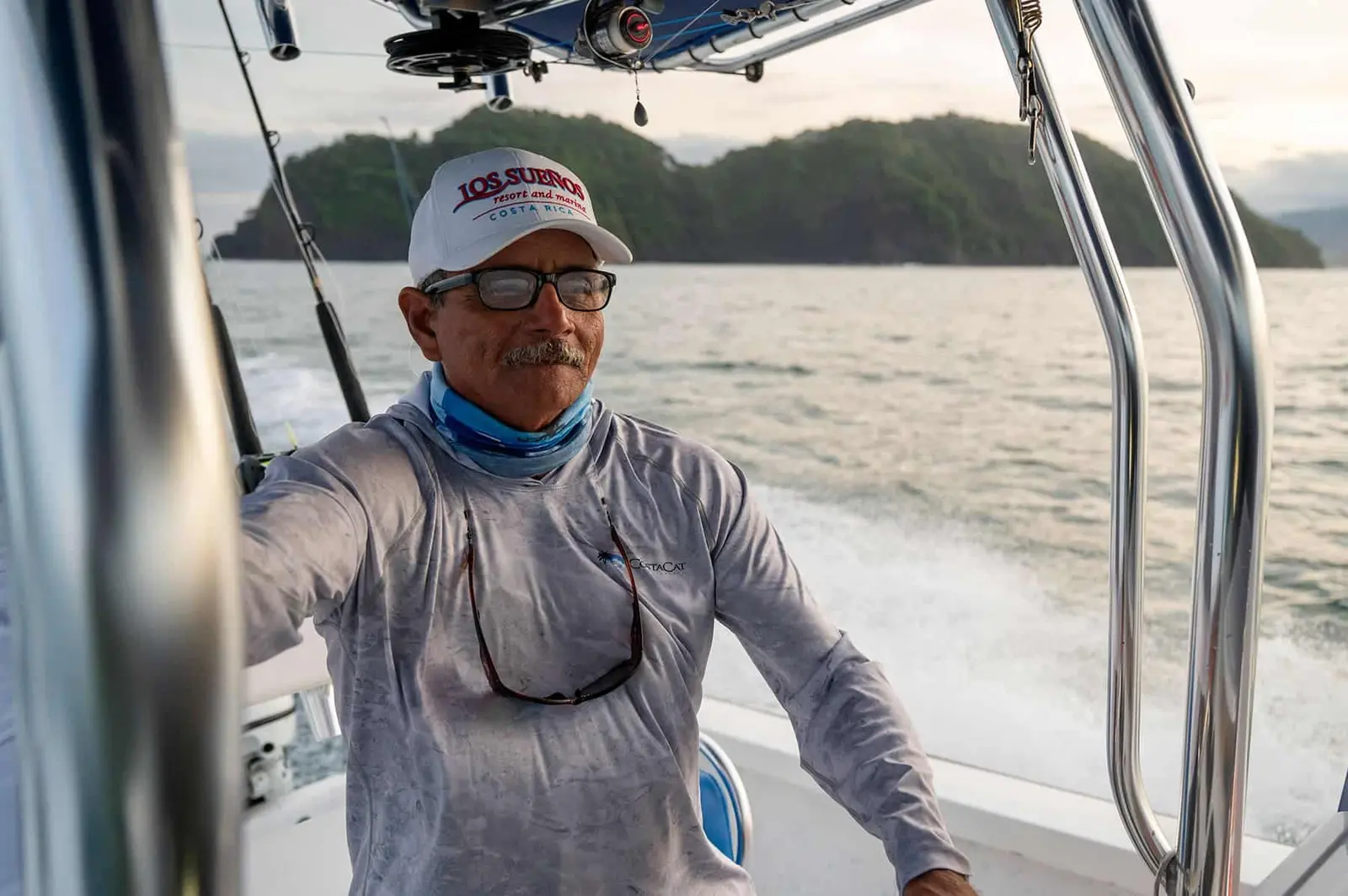 Captain in a cap and sun shirt at the helm of a center-console boat, a green island behind