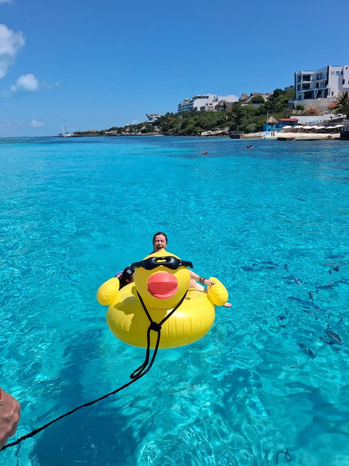 Guest floating on a yellow duck inflatable in clear blue water off Cancún, a rocky shore with houses behind
