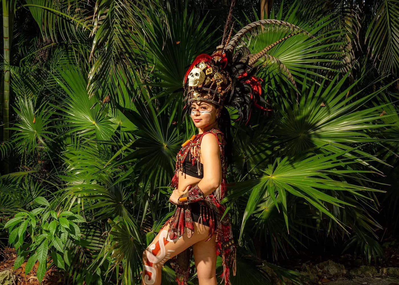Woman in a feathered Mayan-style headdress and costume posing among jungle palms near Cancún