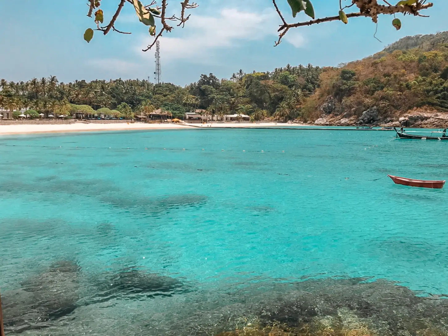 Longtail boat and a small red boat anchored in a clear turquoise bay off a palm-backed Racha beach