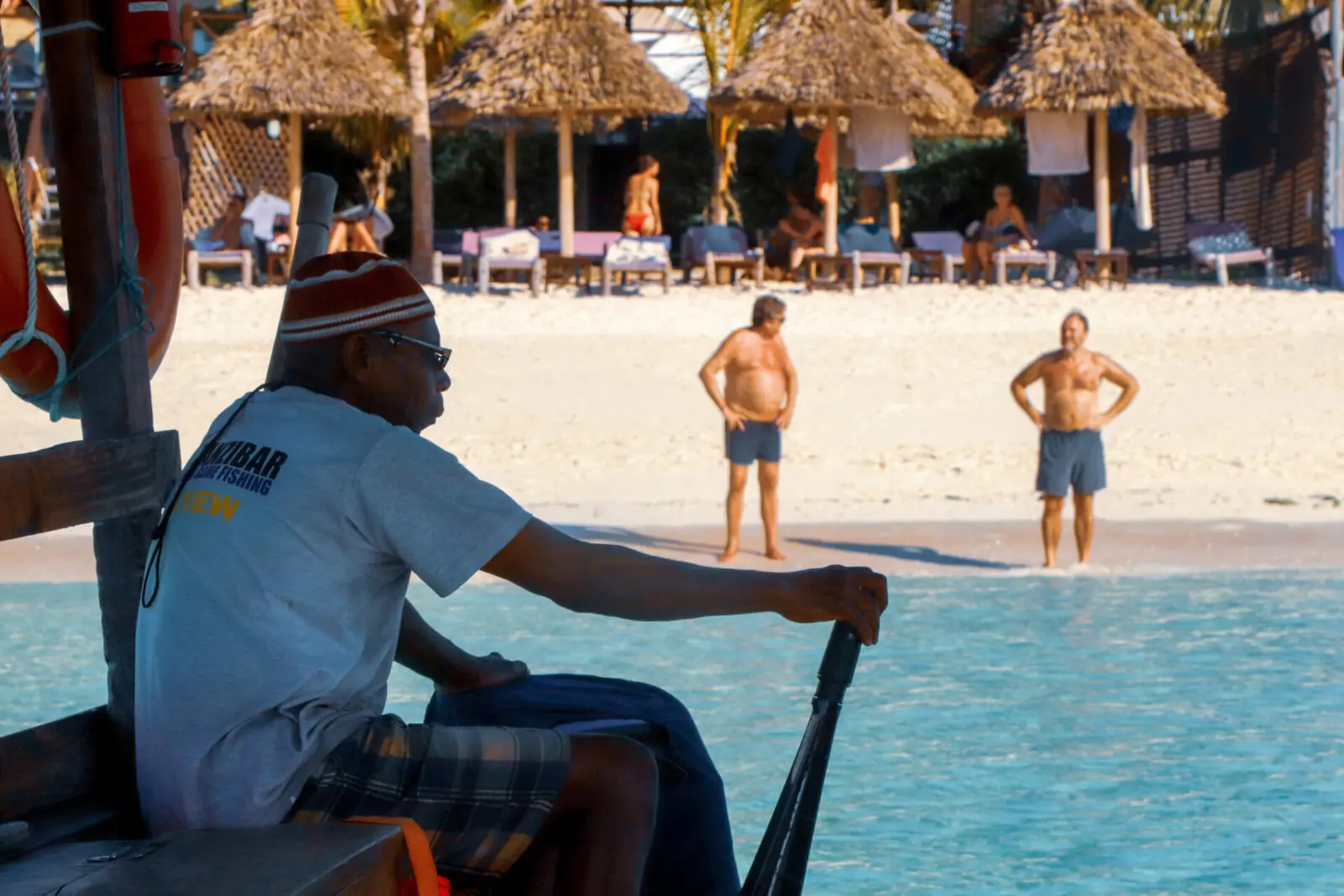 Captain in a knit cap steering a dhow by its tiller past a Zanzibar beach lined with thatched umbrellas