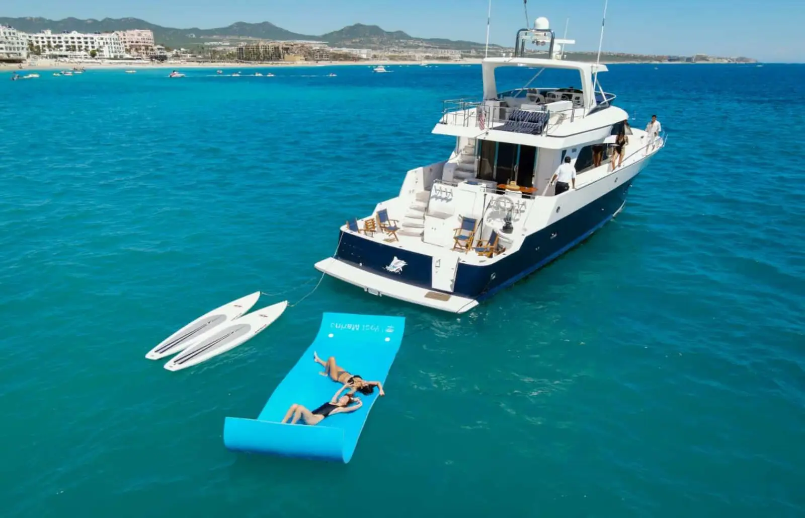 Two guests lounge on a floating mat beside paddleboards behind the motor yacht anchored off Cabo San Lucas