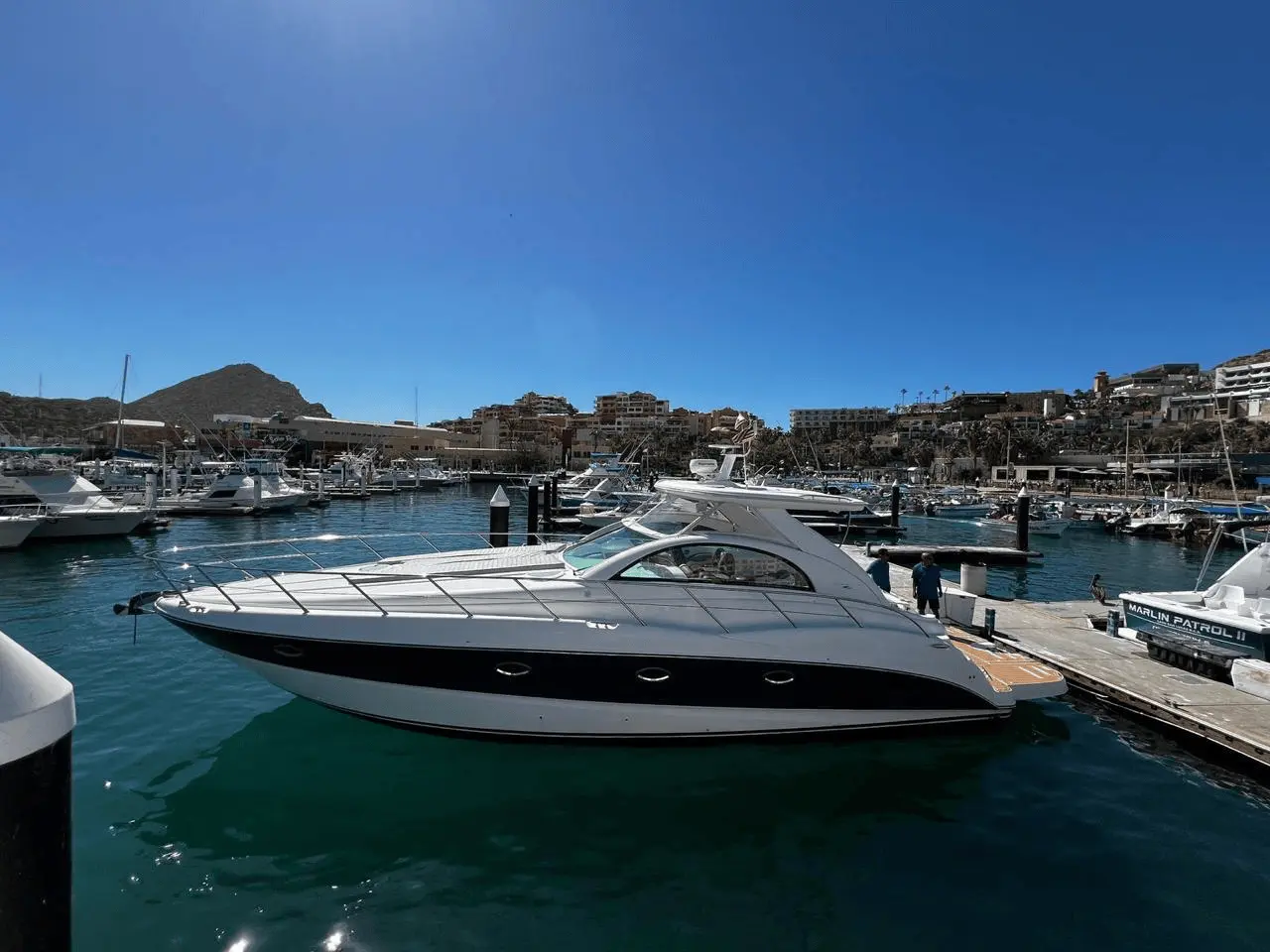 Sleek white motor yacht moored at a marina dock with Cabo San Lucas buildings and hills behind