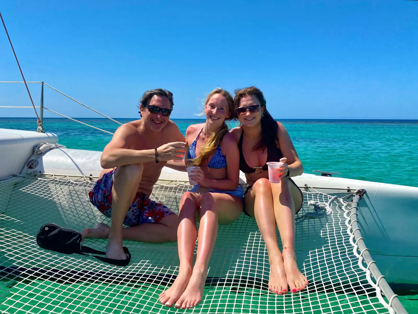 Man and two women with drinks sitting on the netting of a sailing catamaran over turquoise water