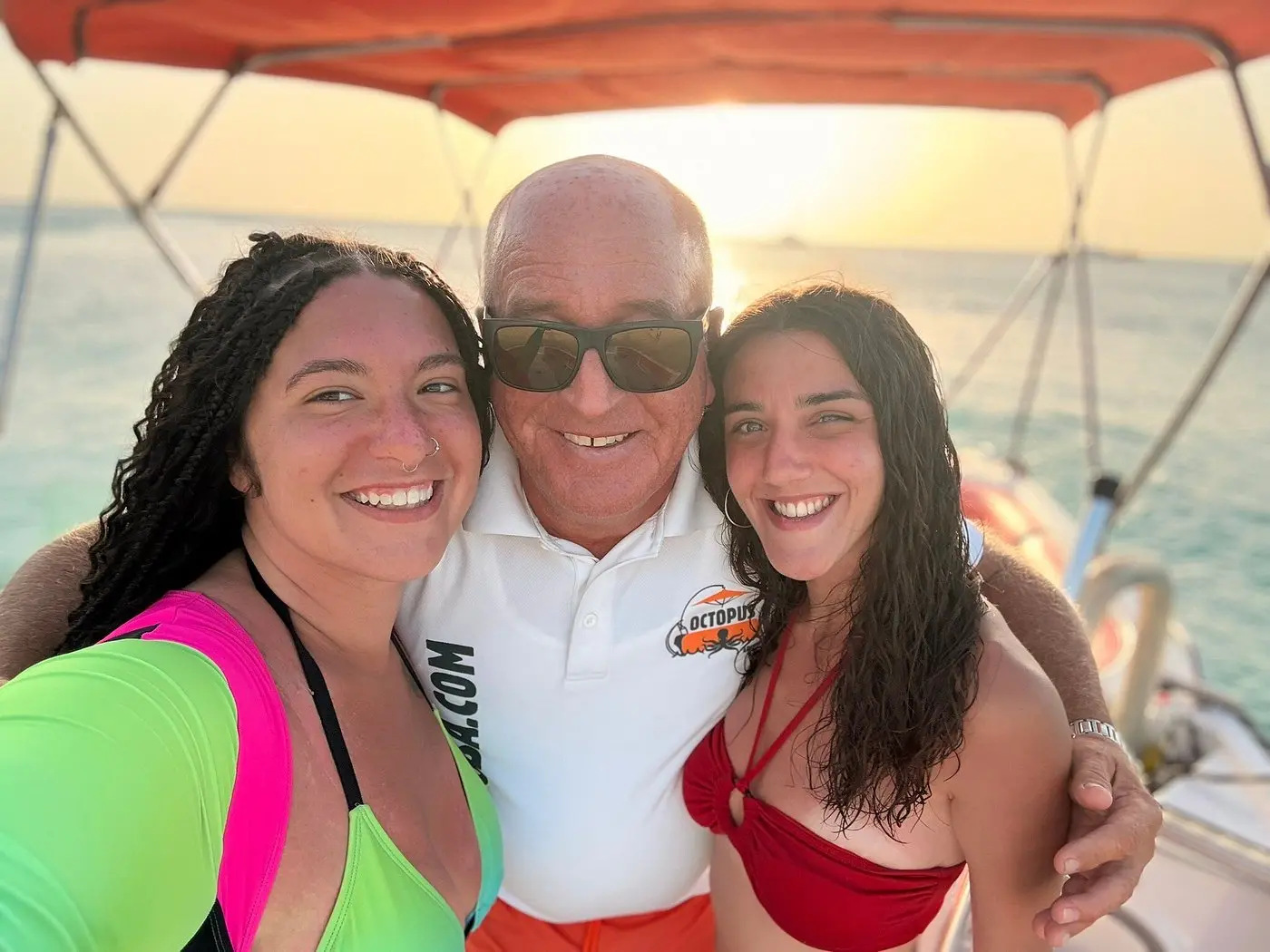 Two women taking a selfie with a crew member under the deck boat's orange canopy at sunset