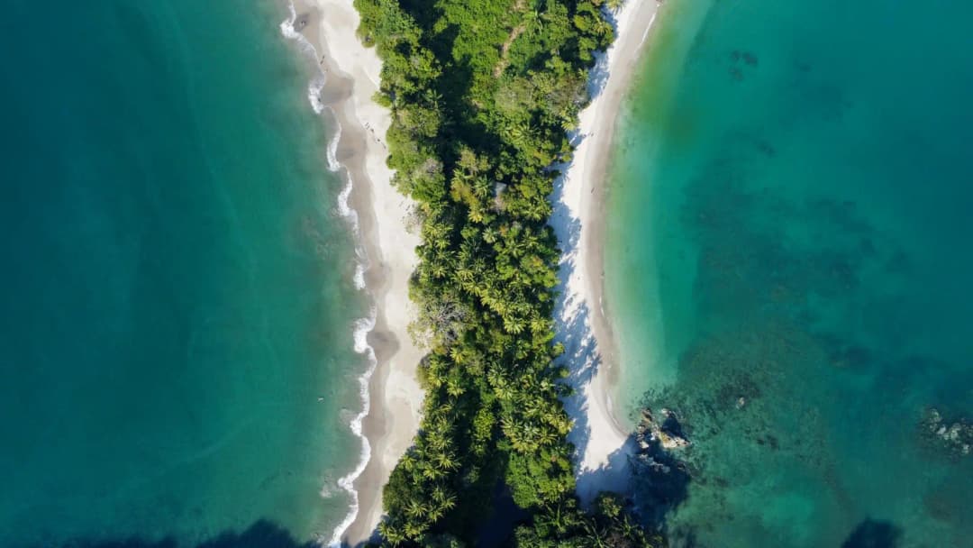 Aerial view of a lush green peninsula separating two turquoise beaches with white sand.

