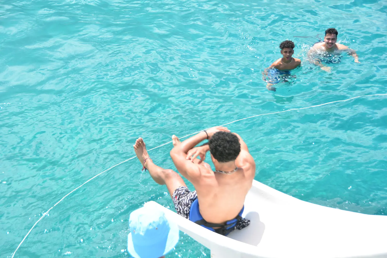 Man riding the catamaran's waterslide toward two swimmers in turquoise water