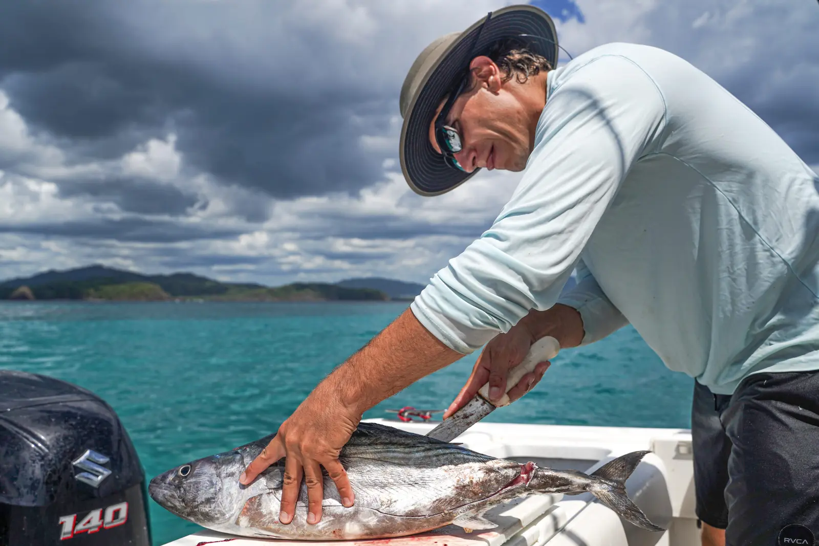 A man in a sun hat fillets a fresh tuna on the stern of the fishing boat, green coast behind