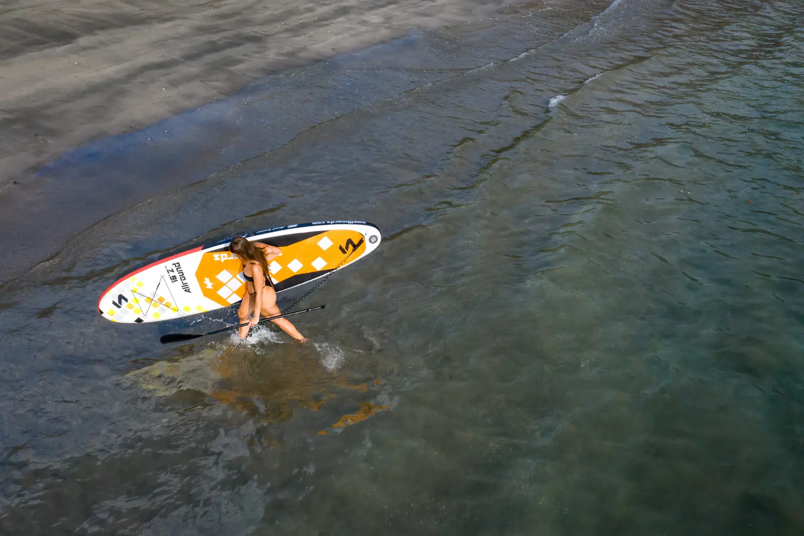 A woman wades through clear shallows with a stand-up paddleboard on the Guanacaste coast