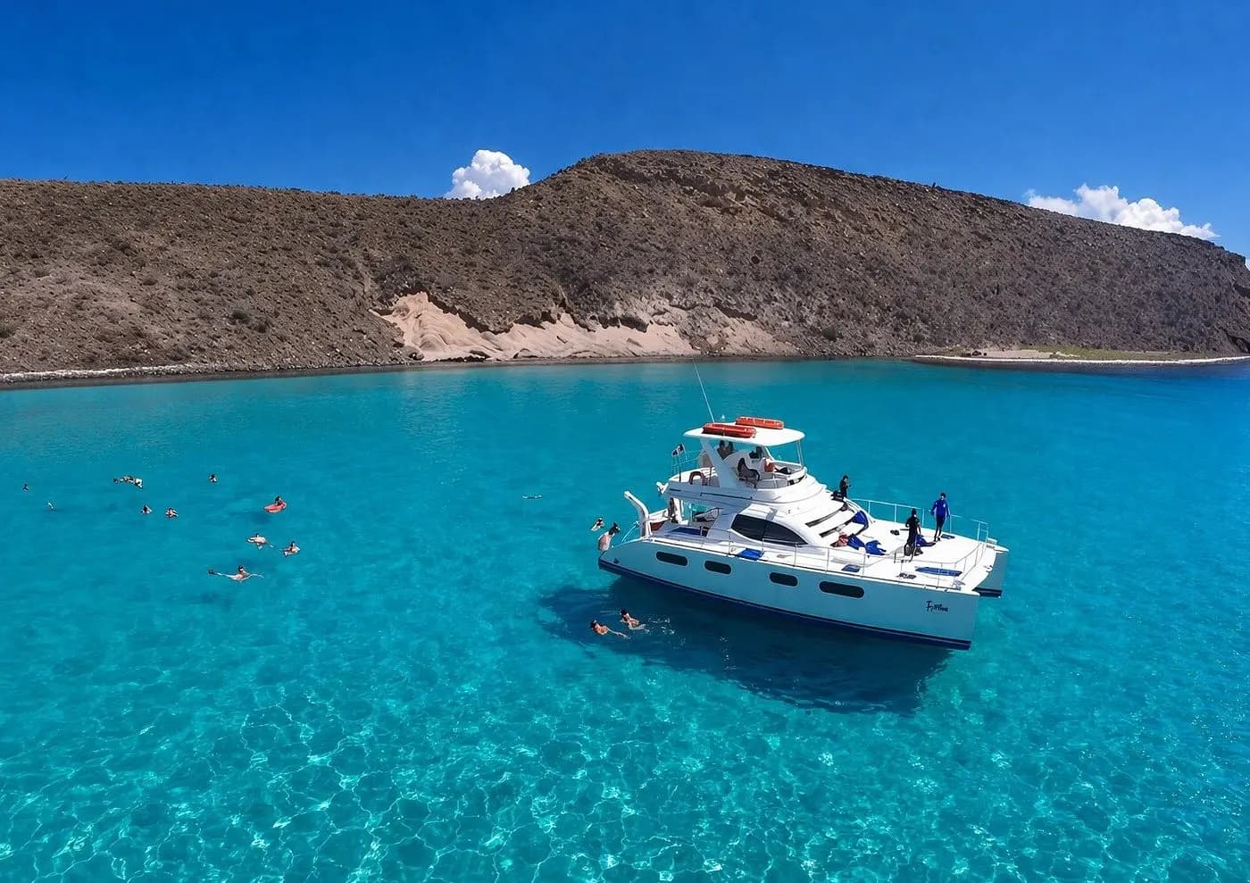 Catamaran anchored in turquoise shallows below a desert hill near La Paz as guests swim nearby