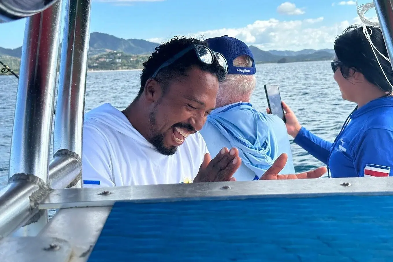 Guests sharing a laugh aboard the fishing boat while one looks at a phone, green hills beyond