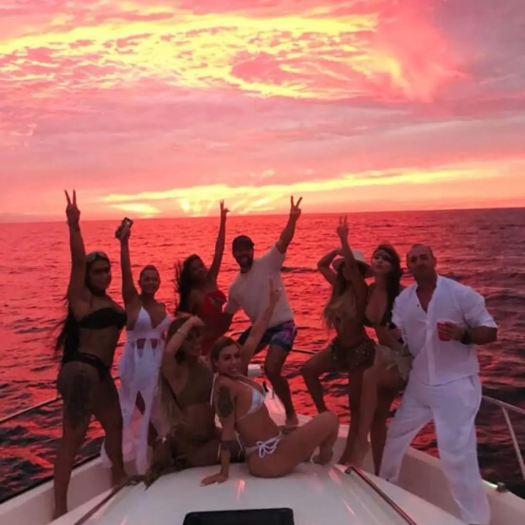 Group of friends cheering on a yacht's bow under a red and pink sunset sky off Cabo San Lucas