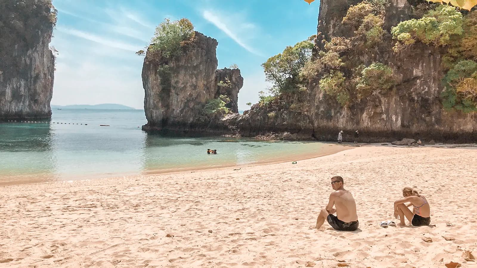 Two guests sitting on a white-sand beach facing limestone sea stacks in Phang Nga Bay