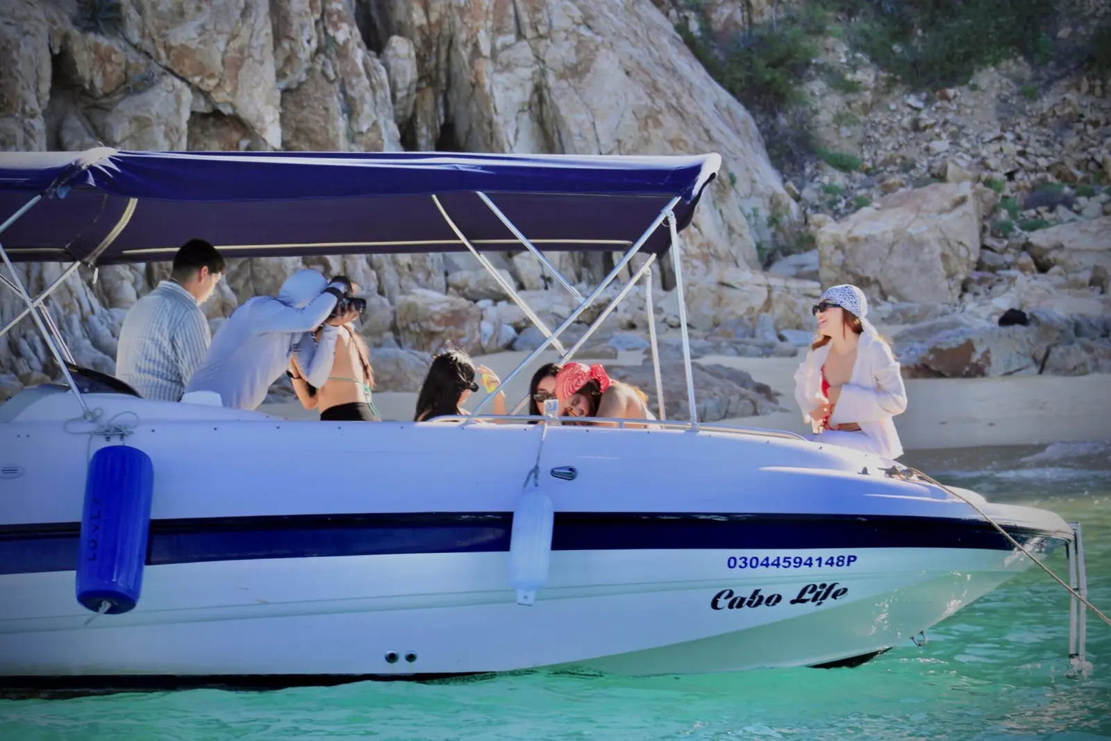Guests seated under the navy canopy of a small yacht moored beside granite boulders in Los Cabos