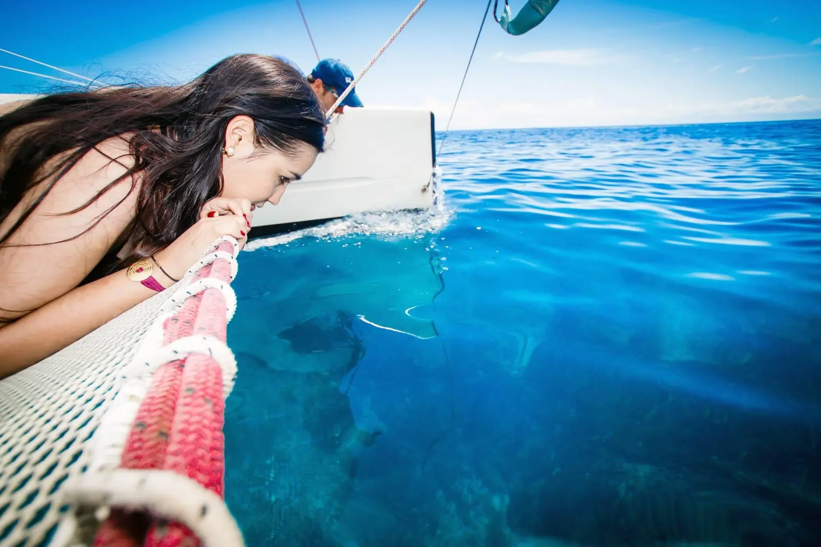 Woman lying on the catamaran's netting, peering down into the clear blue water as the boat sails