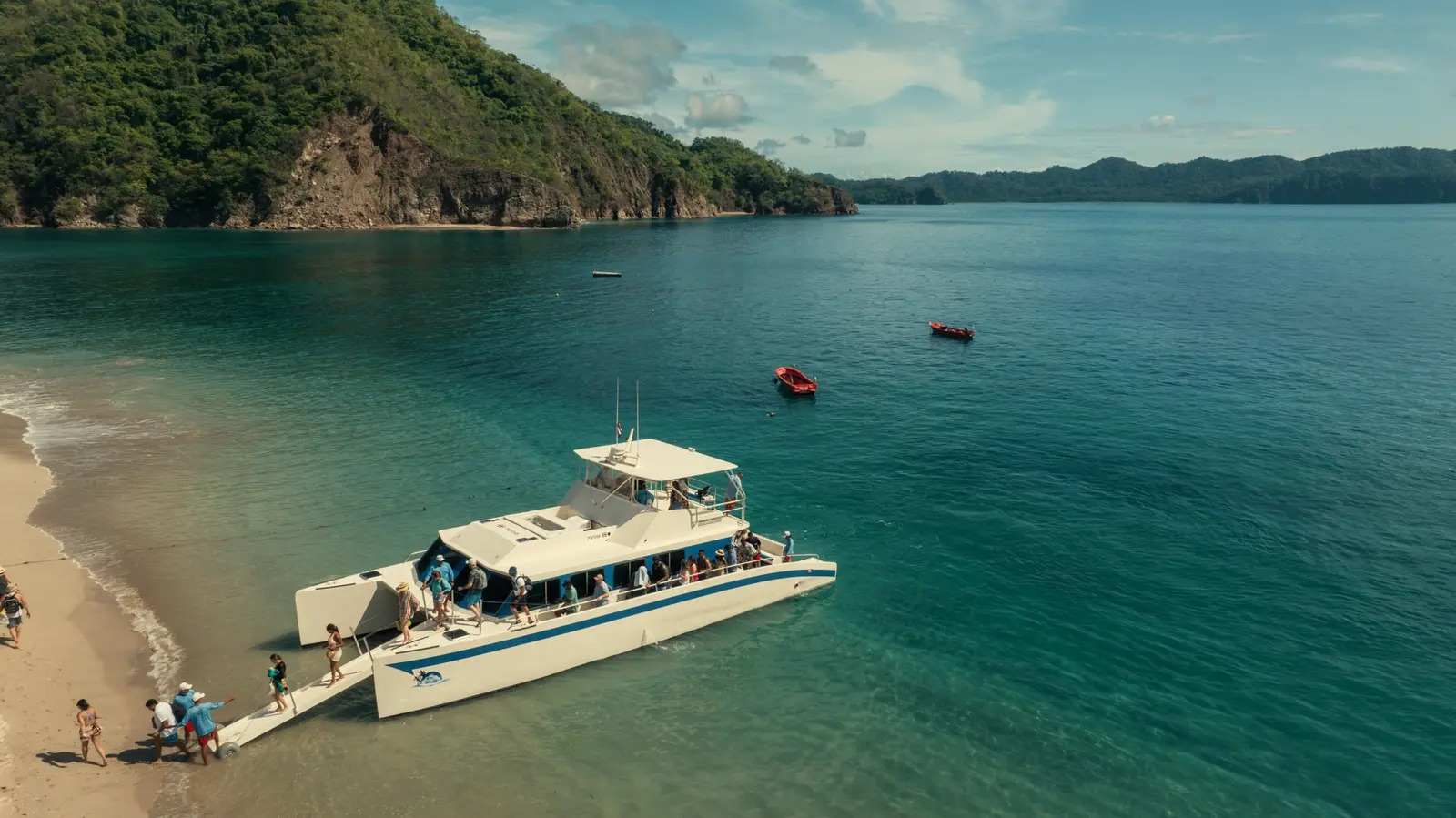 High aerial view of the catamaran at the water's edge in a turquoise Costa Rica bay, small red boats nearby