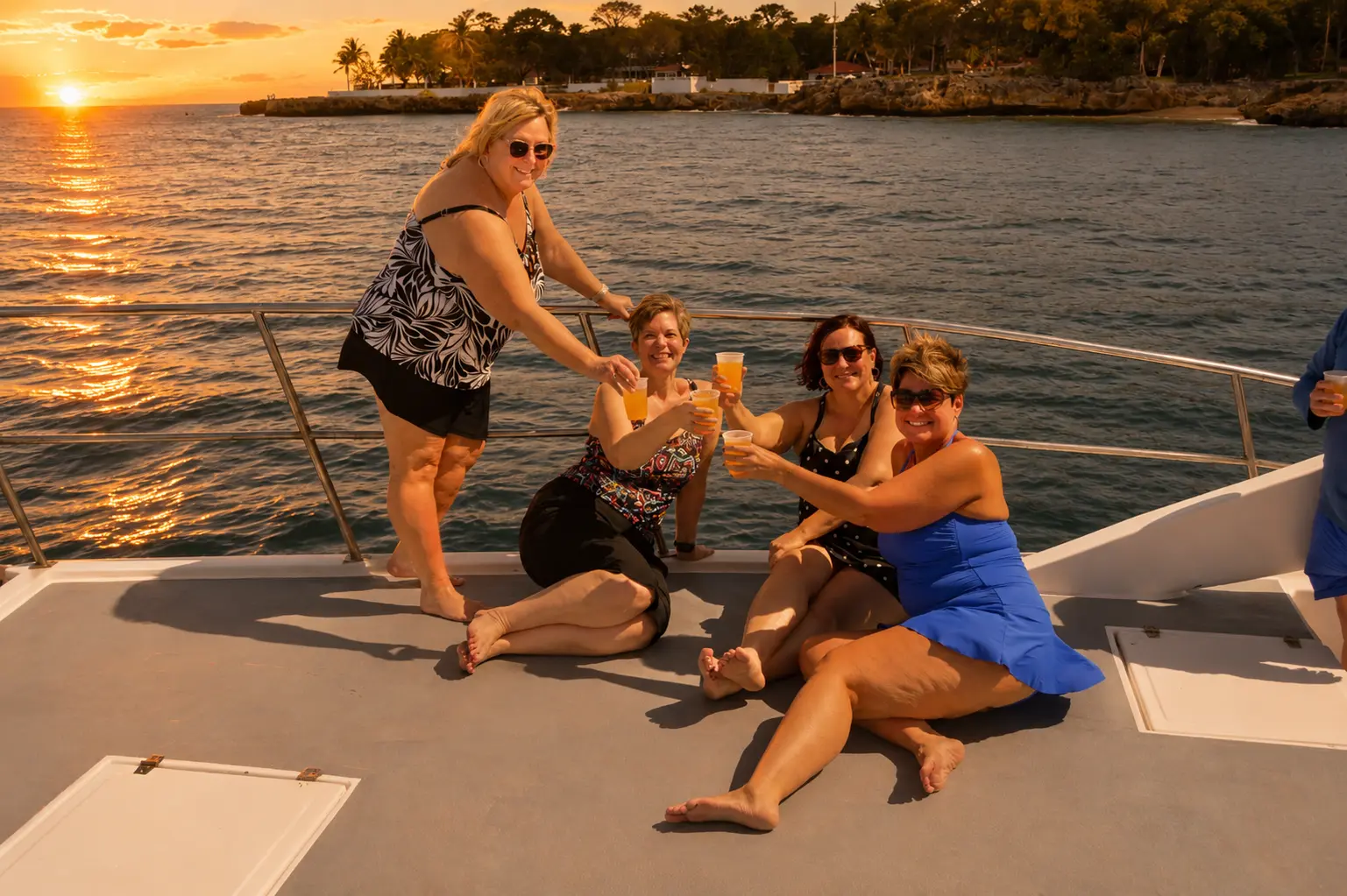 Four women posing with drinks on a catamaran's foredeck at sunset off the Puerto Plata coast