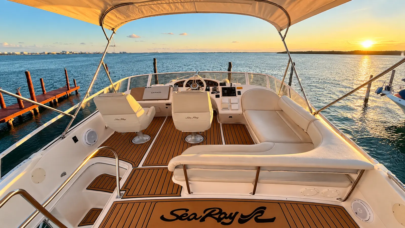 Flybridge deck with helm seats, a white lounge and teak flooring as the sun sets over a Cancún marina