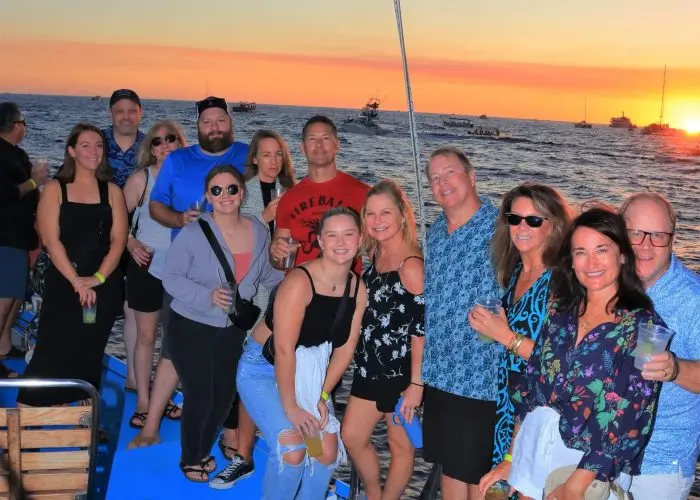 Group of adults with drinks gathered on a catamaran deck as the sun sets off Cabo San Lucas