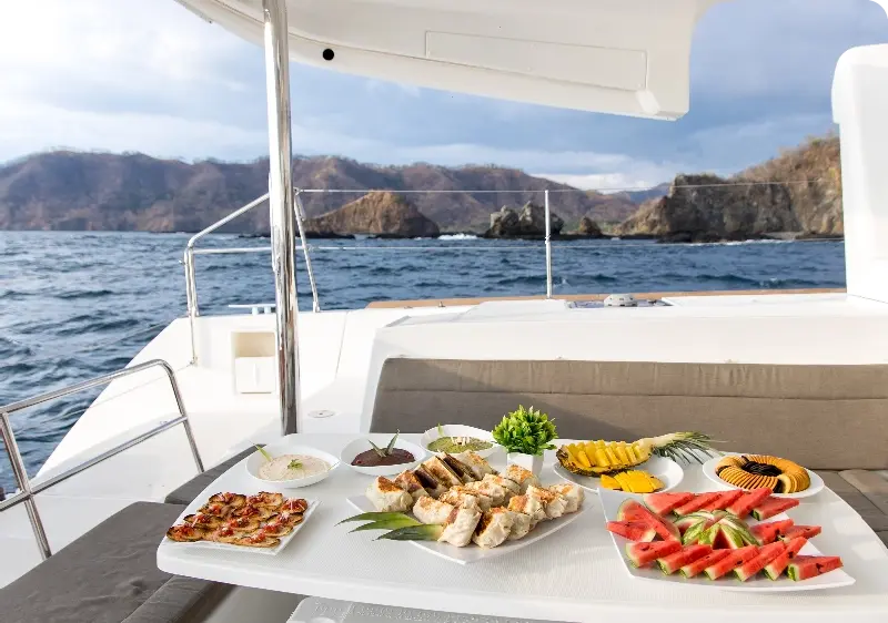 Lunch spread of watermelon, wraps and dips on the catamaran's shaded aft table, rocky islets beyond