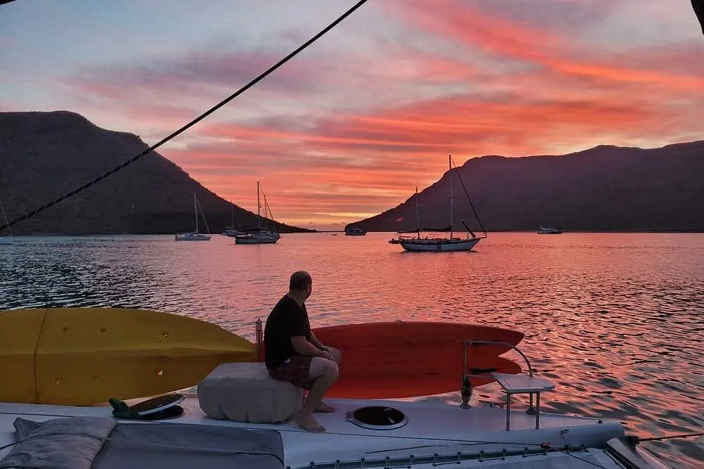 A man on a sailboat deck watching a pink sunset over boats anchored in a calm bay near La Paz