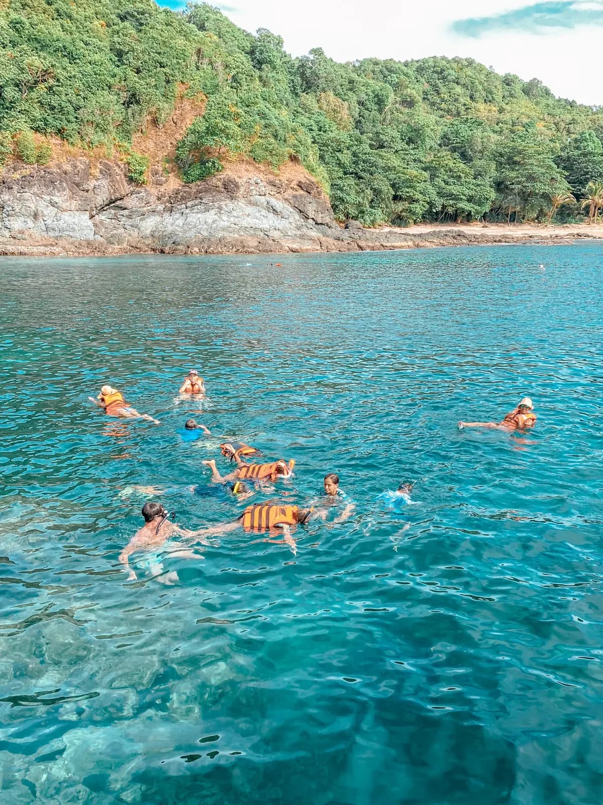 Snorkelers floating in clear blue water off the rocky, forested shore of a Khai island