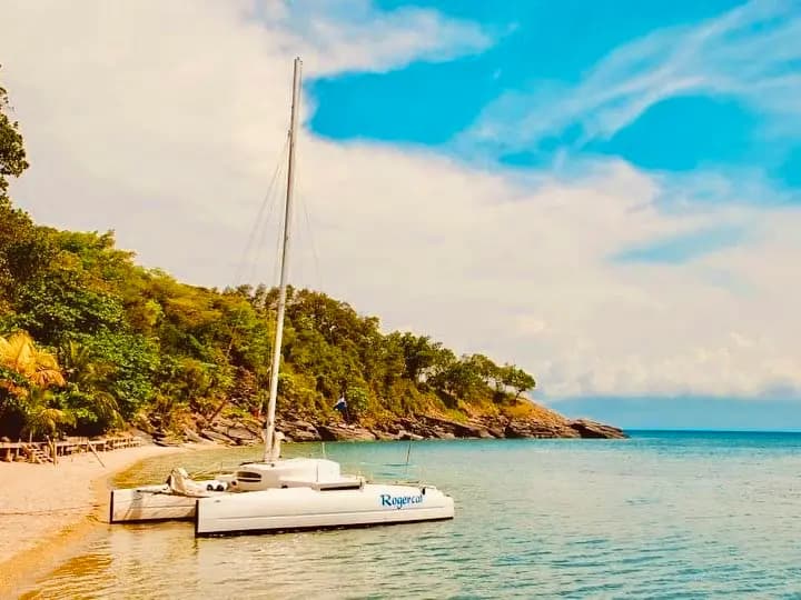 Sailing catamaran anchored in clear shallows by a sandy beach and wooded headland on Roatan