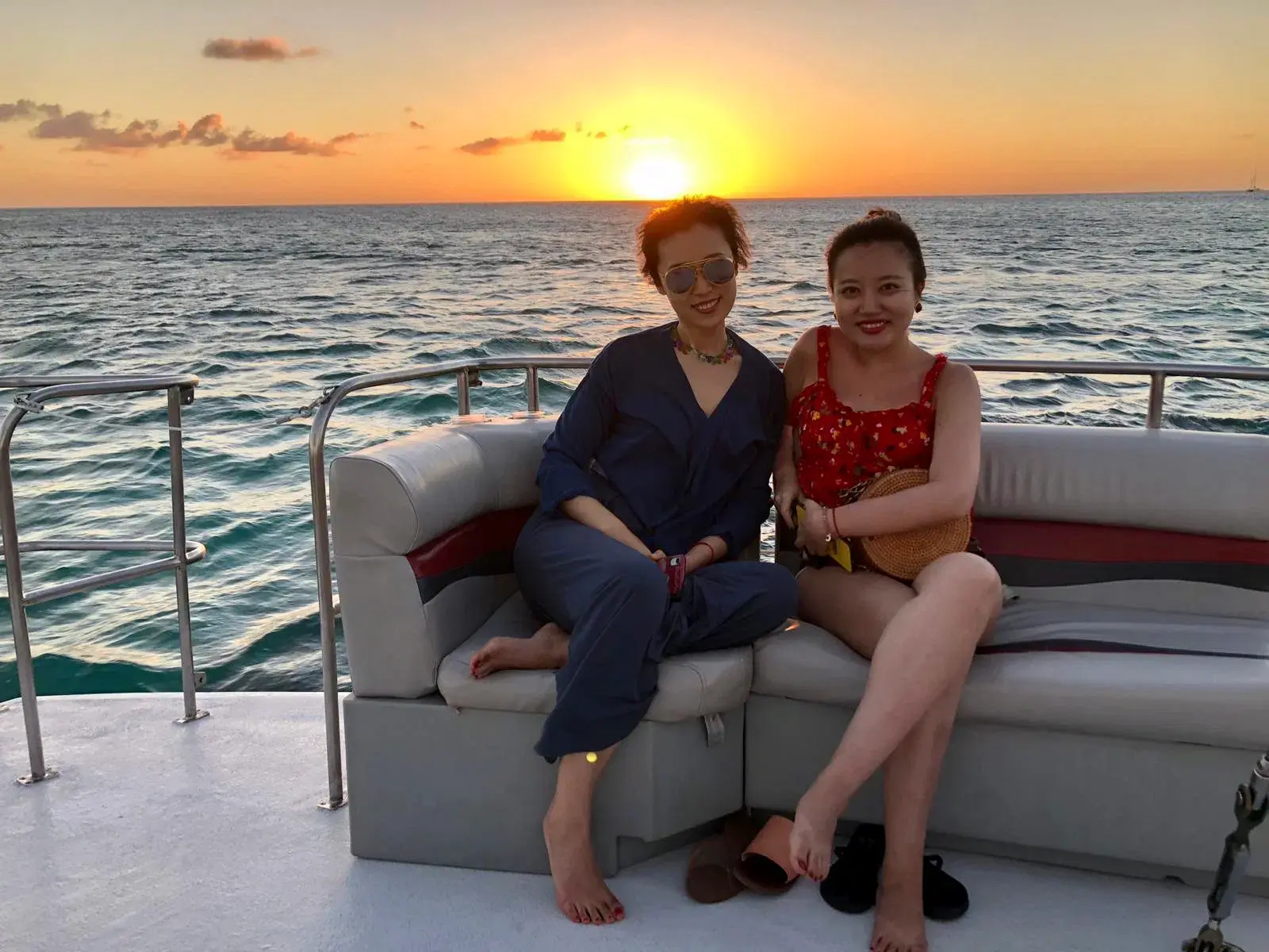 Two women relaxing on a cushioned lounge sofa aboard a catamaran as the sun sets