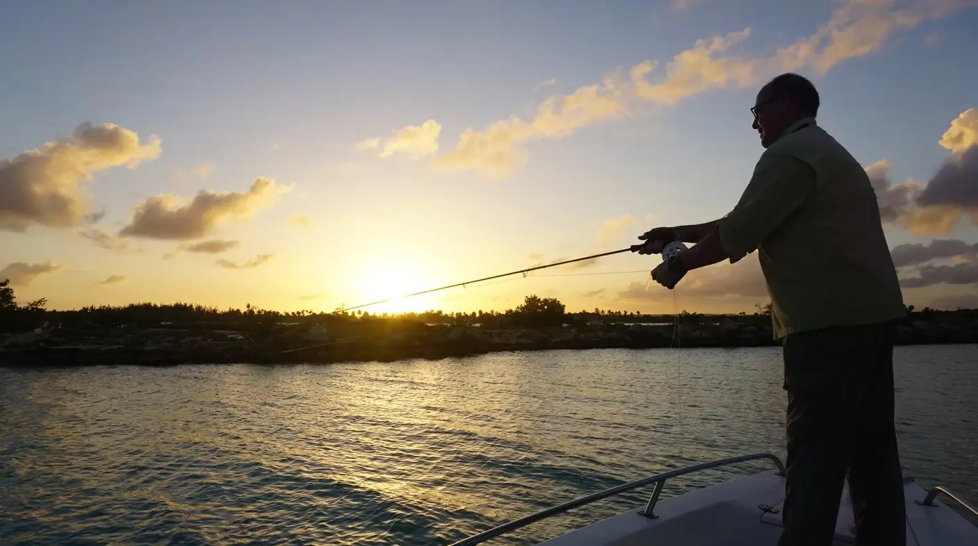Silhouetted angler casting from the bow of a fishing boat at sunset over a tree-lined Punta Cana shore