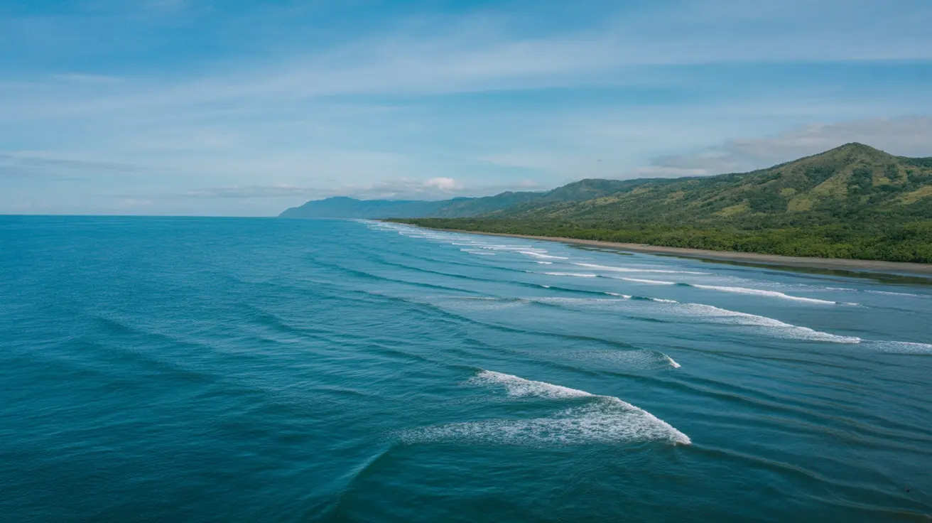 Aerial view of waves rolling toward a long dark-sand beach backed by green forested hills in Costa Rica