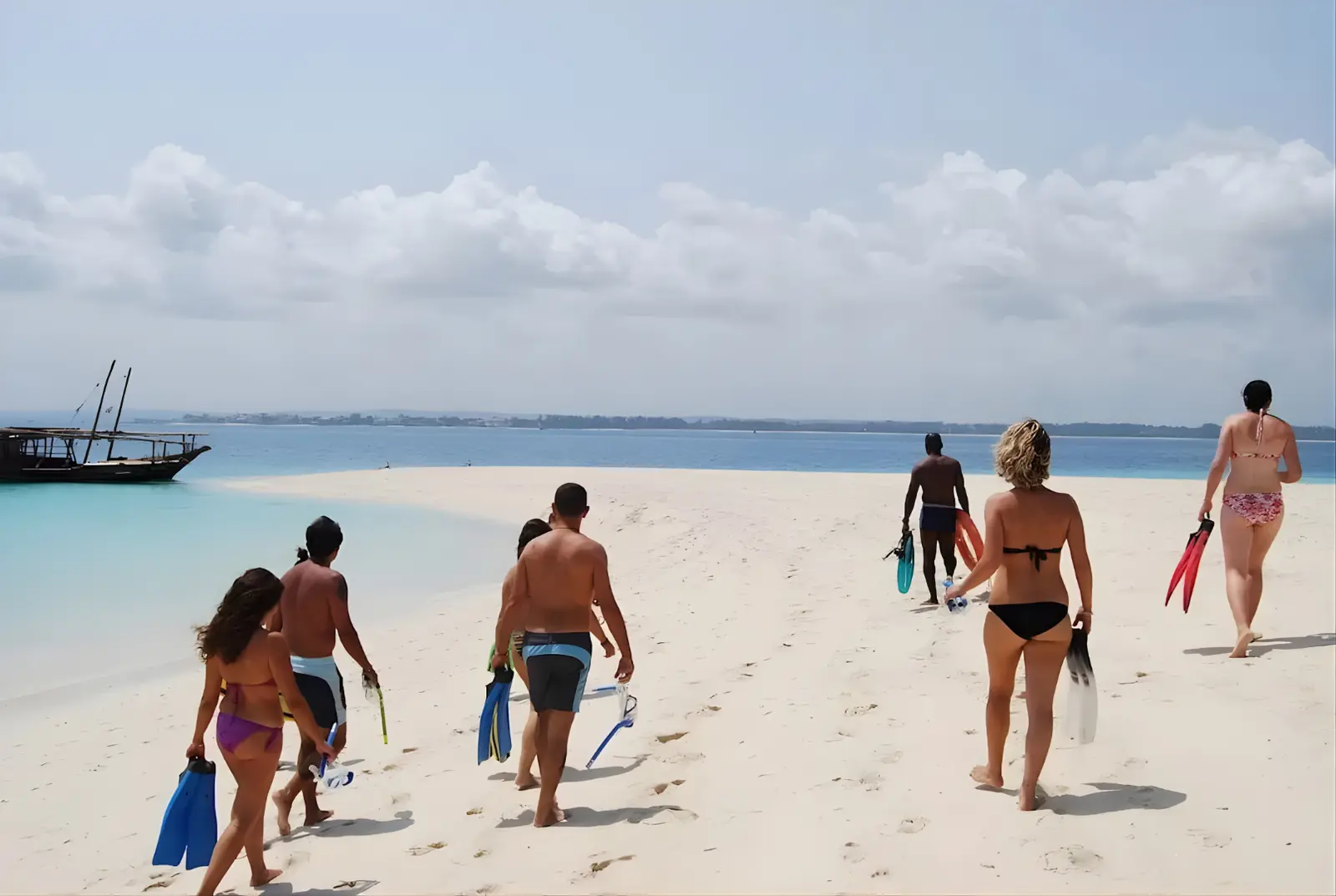 Guests carrying snorkel fins across Nakupenda Sandbank toward the water, a dhow moored nearby