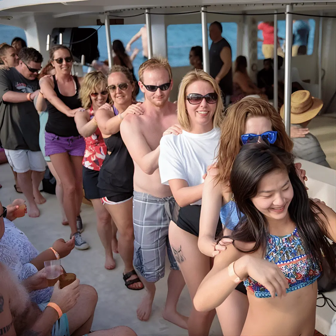 Guests forming a conga line on the covered deck of a catamaran, hands on each other's shoulders