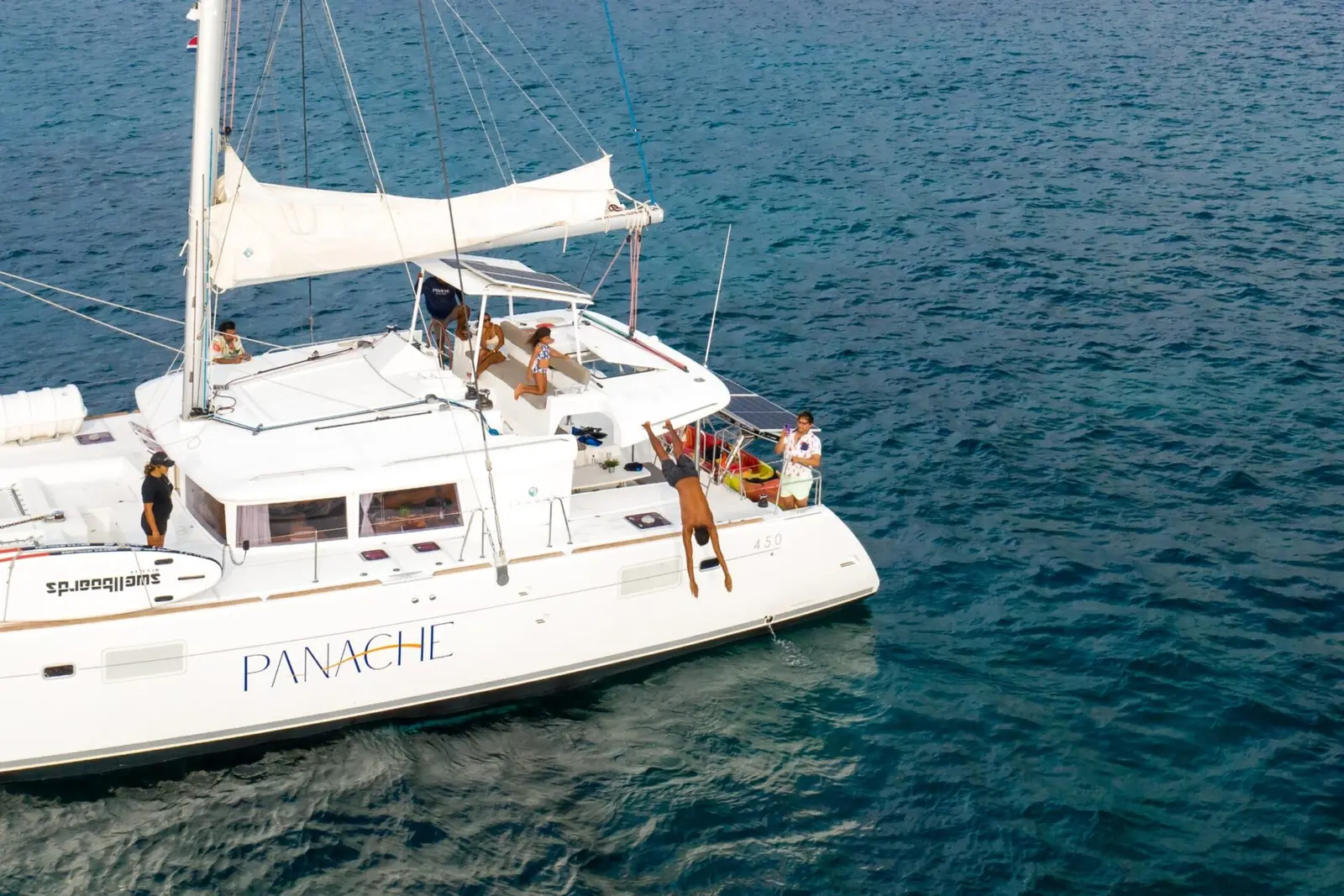 Guest leaping off the side of a sailing catamaran into deep blue water as others relax on deck