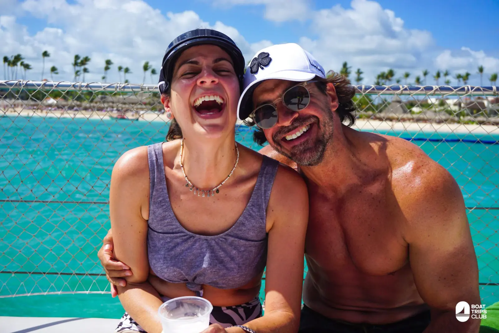 Laughing couple in caps on a catamaran deck, turquoise water and the palm-lined Bavaro shore behind