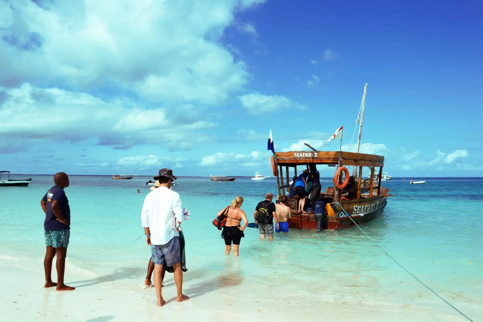 Guests wading through shallow turquoise water to board a wooden dhow from a Zanzibar beach