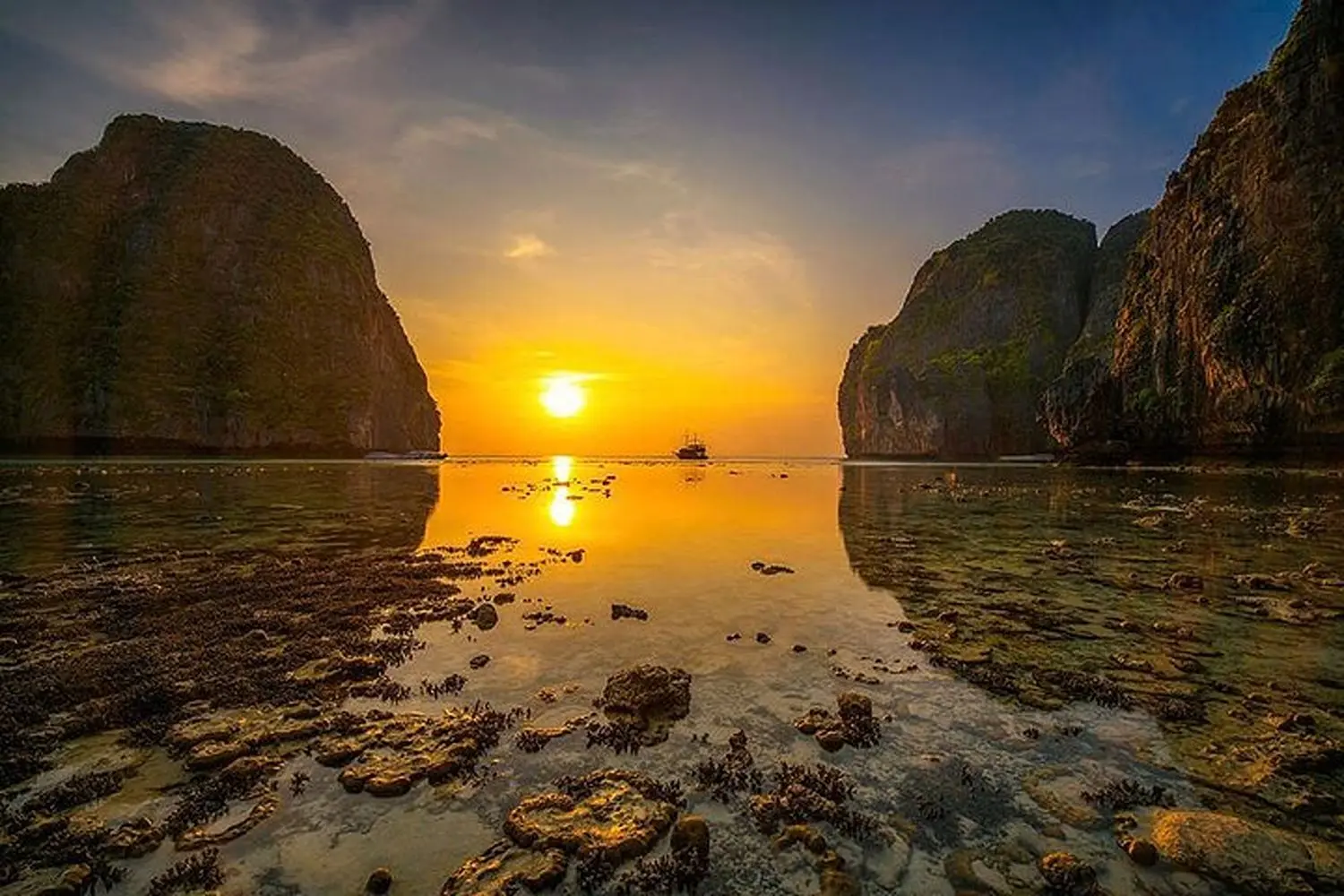 Sun setting between two limestone cliffs, reflected in a rocky low-tide bay in Thailand