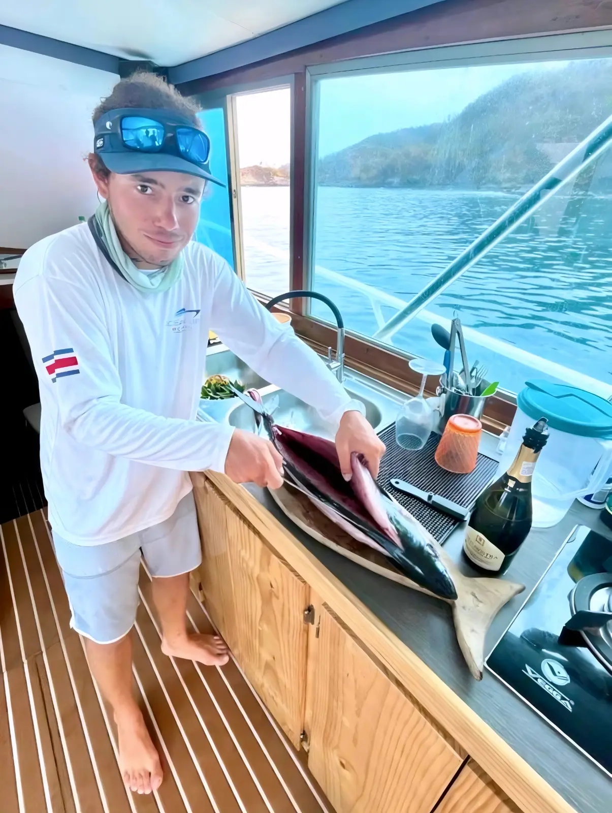 A man fillets fish on a cutting board in the yacht's galley, green hills outside the window