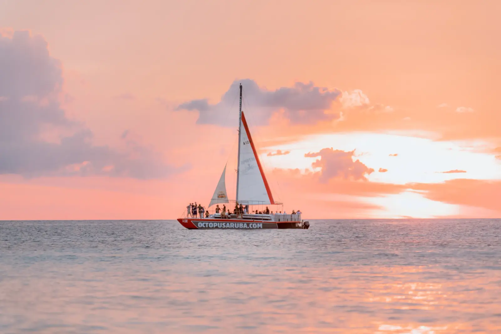 Sailing catamaran with guests aboard cruising calm water under a pink sunset sky off Aruba