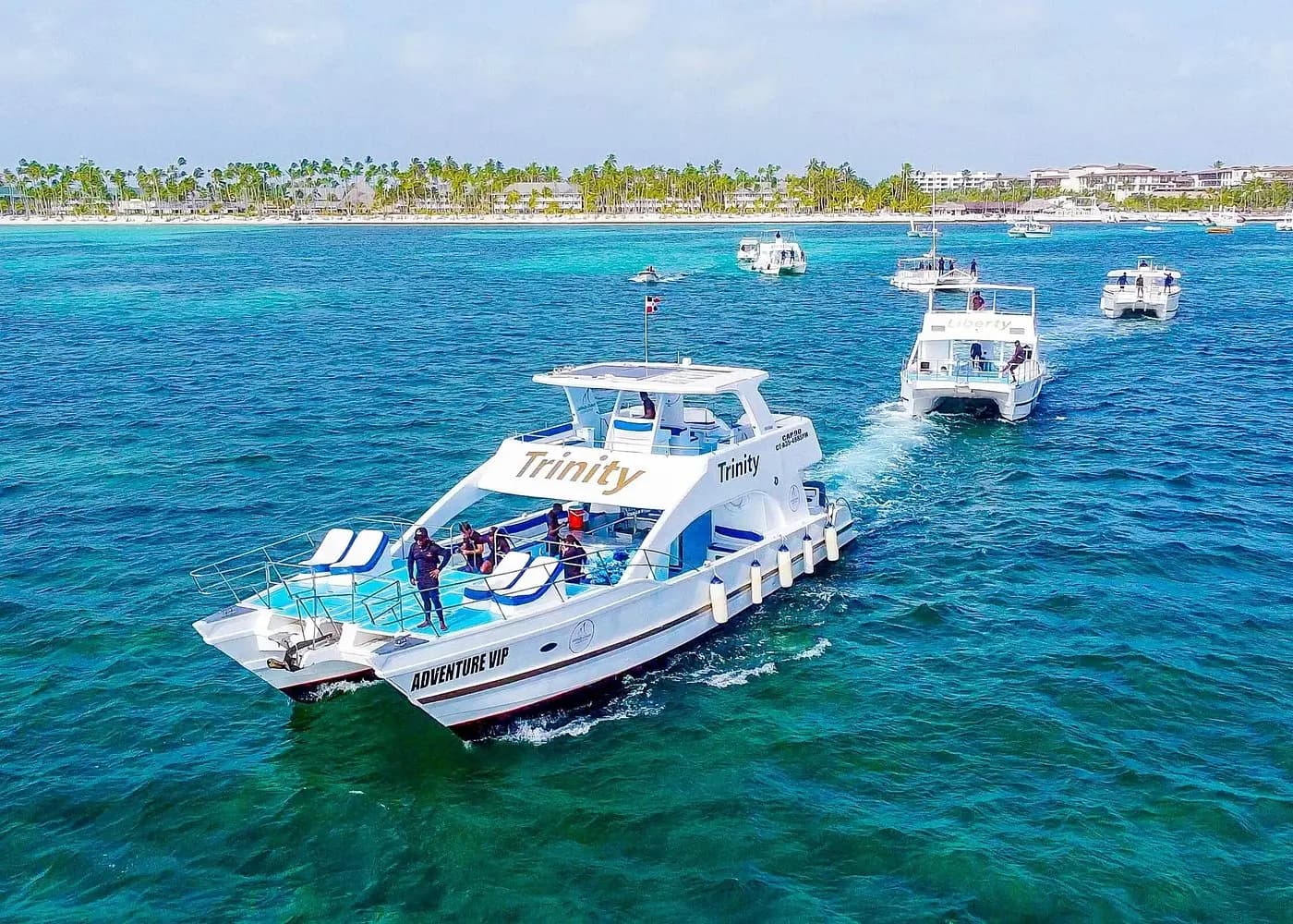 Aerial view of a white catamaran leading other boats across blue water off the palm-lined Punta Cana beach