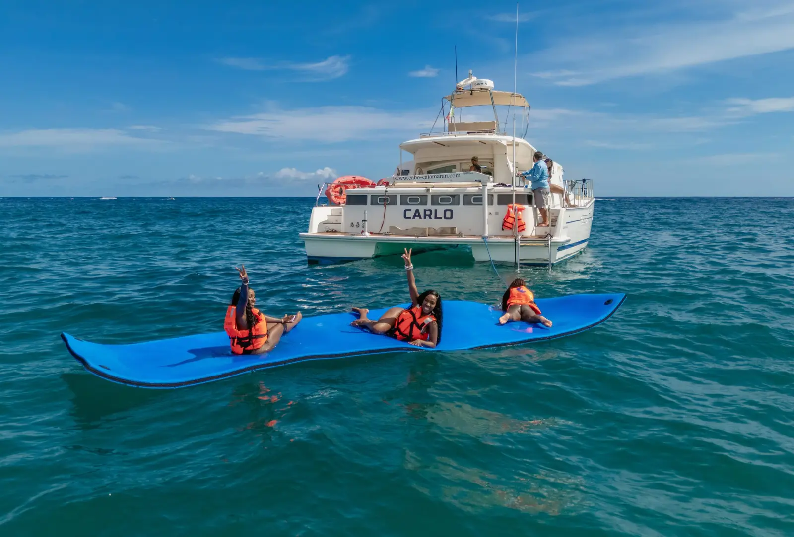 Guests in life vests waving from a blue floating mat beside a power catamaran anchored off Cabo San Lucas