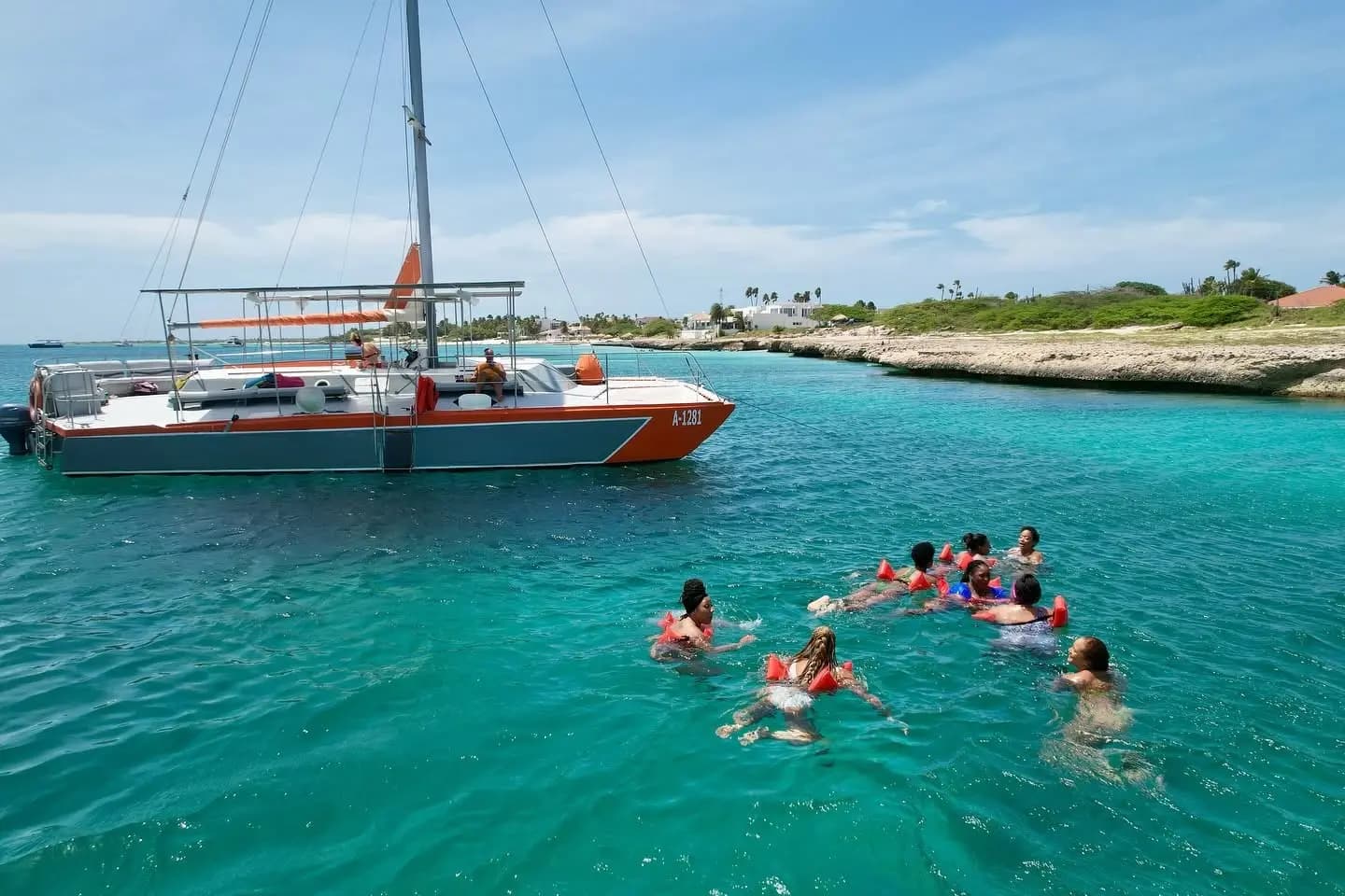 Snorkelers in orange vests swimming beside an anchored sailing catamaran off Aruba's rocky coast