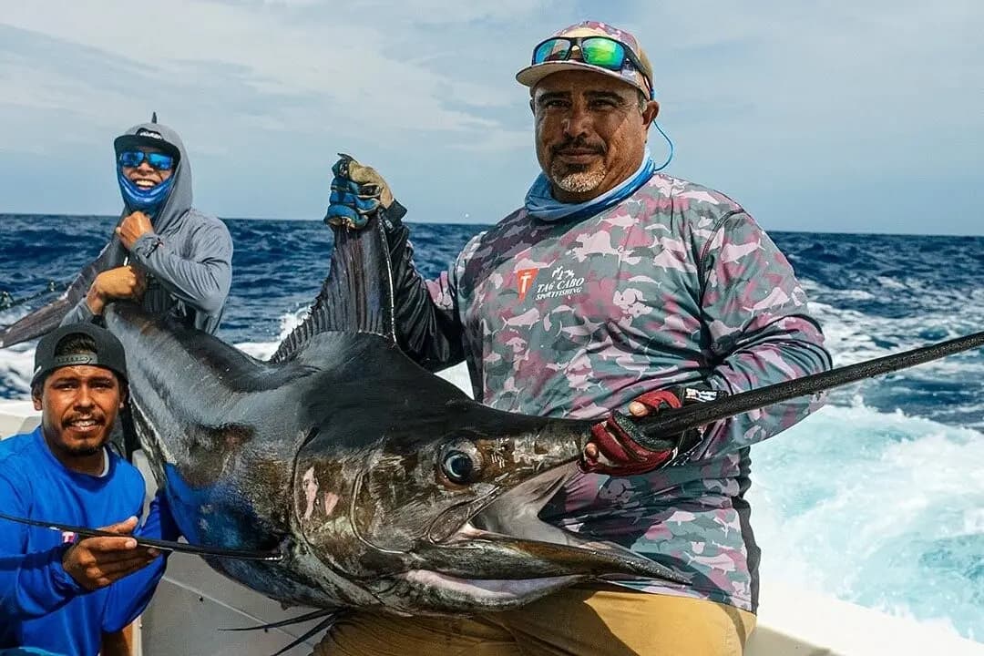 Crew members lift a large marlin by its bill at the side of the boat with whitewater behind
